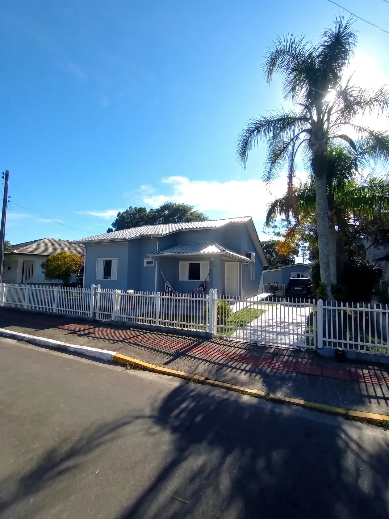 A casa vista da rua, com a cerca branca, a palmeira e a calçada em frente.