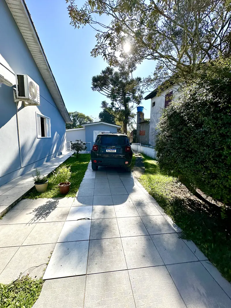 Corredor lateral da casa com carro estacionado e uma araucária alta ao lado.