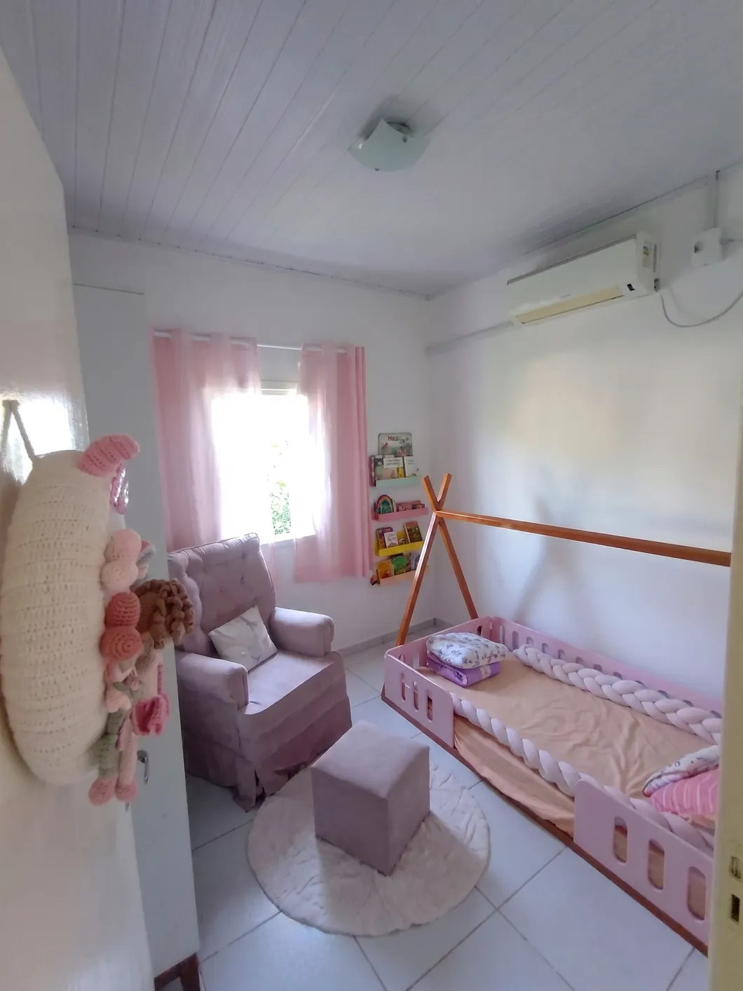 Quarto infantil com cama montessoriana, poltrona de amamentação, prateleira de livros e cortina rosa.